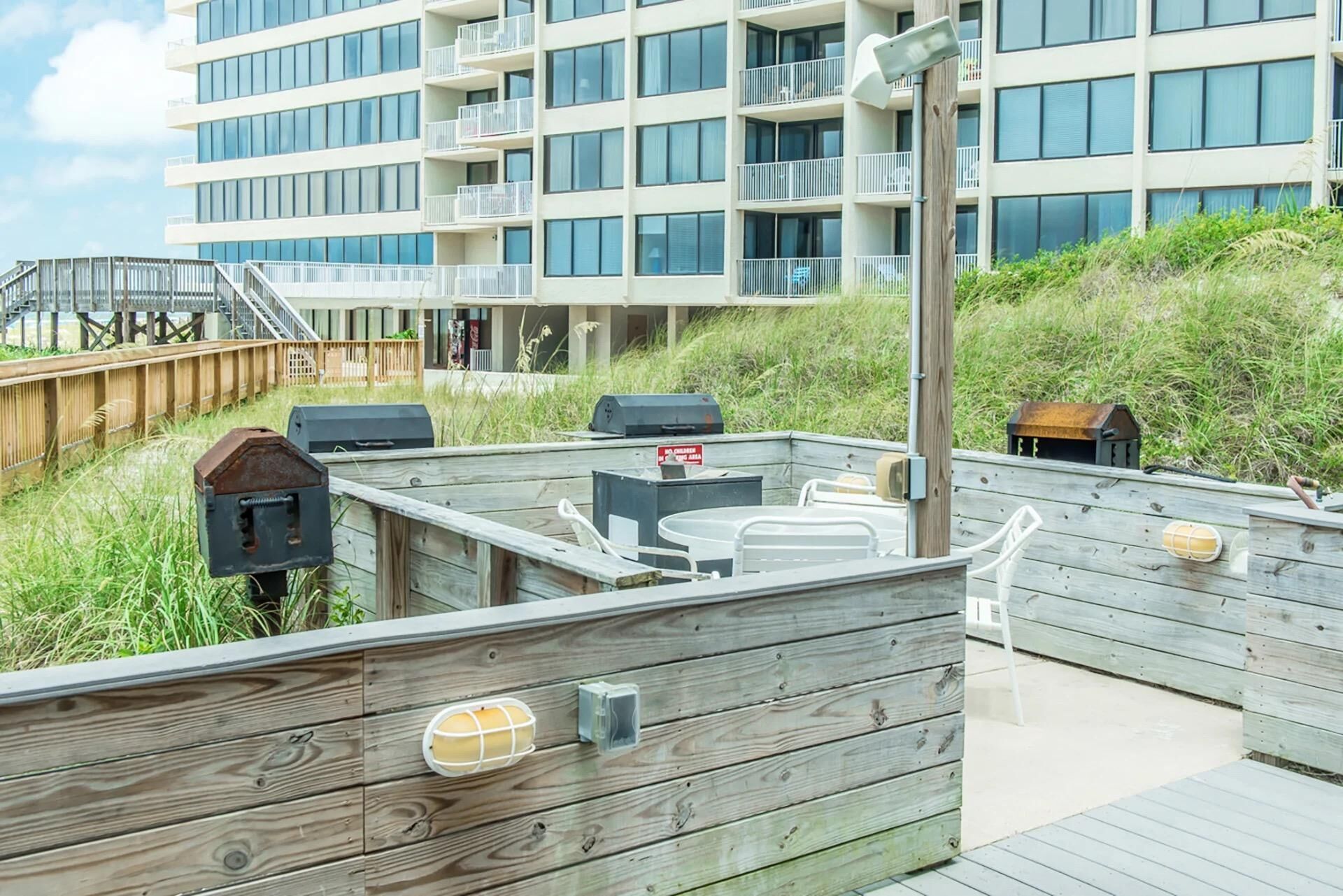 Terraza de madera con mobiliario exterior, una parrilla de barbacoa y vistas a un edificio de varios pisos con balcones.