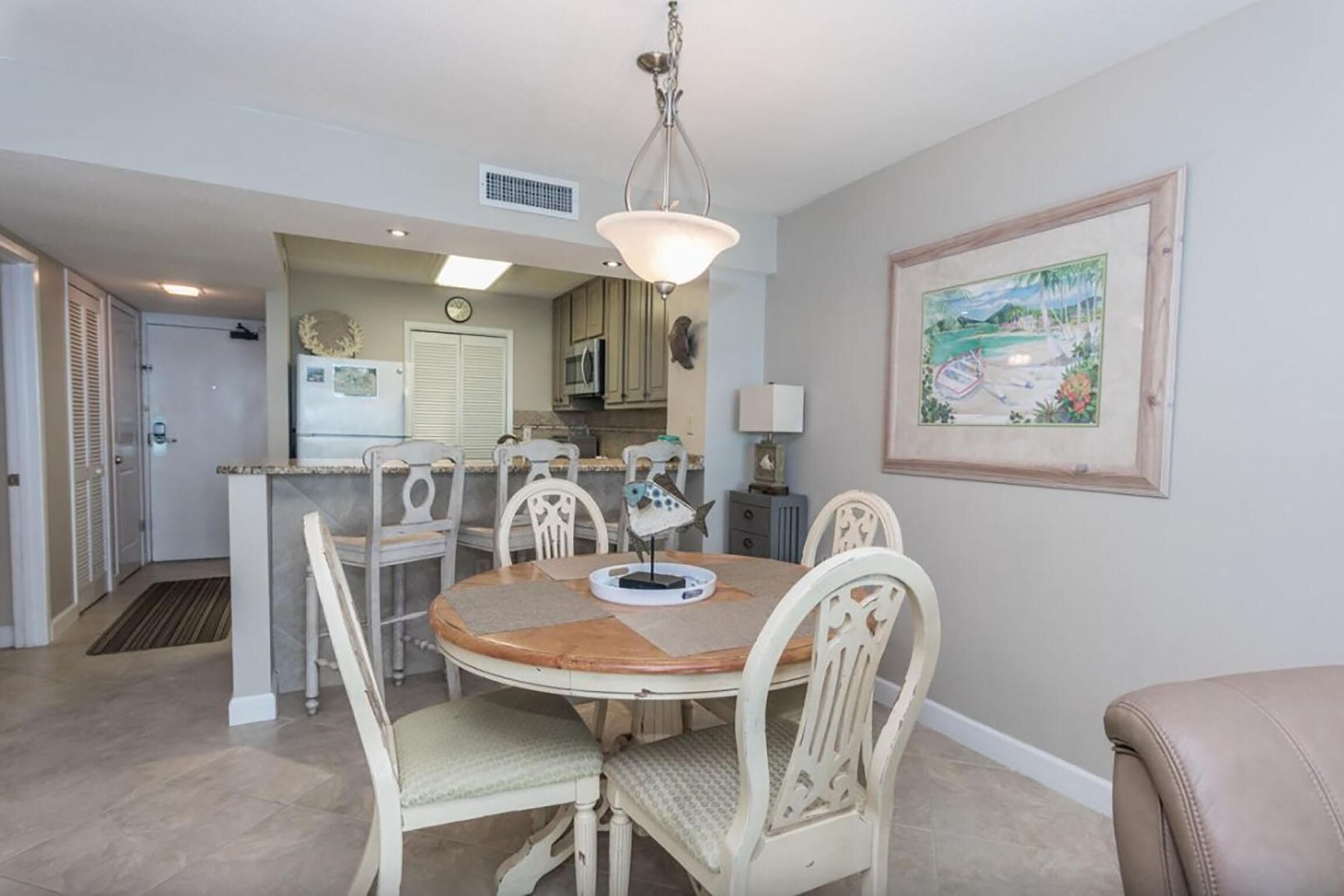 A dining area with a round table, white chairs, and a pendant light. There is a kitchen in the background with cabinets and a microwave.