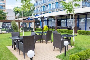 An outdoor dining area with wicker furniture and a covered section, surrounded by greenery and a building with balconies.