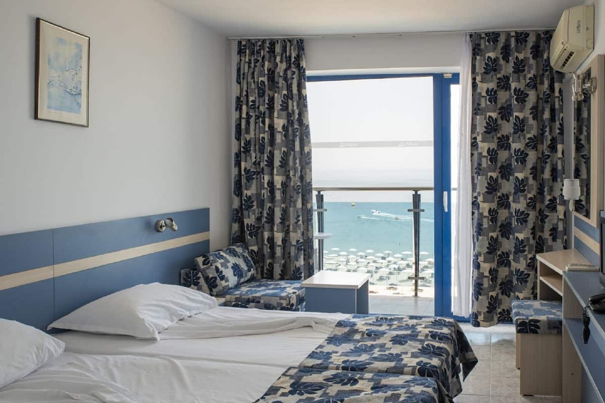 A hotel room with a bed, a seating area, a small table, and a view of the beach and sea.