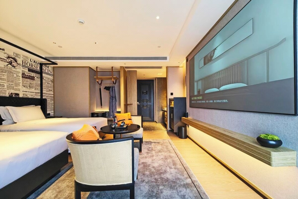 Quarto de hotel moderno com uma cama grande, área de estar com uma mesa, televisão fixada na parede e uma pequena planta em uma prateleira.