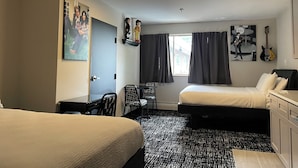 Una camera d'albergo con due letti, una scrivania, una sedia, una finestra con tende, una chitarra e opere d'arte alle pareti, tra cui un poster dei Beatles.