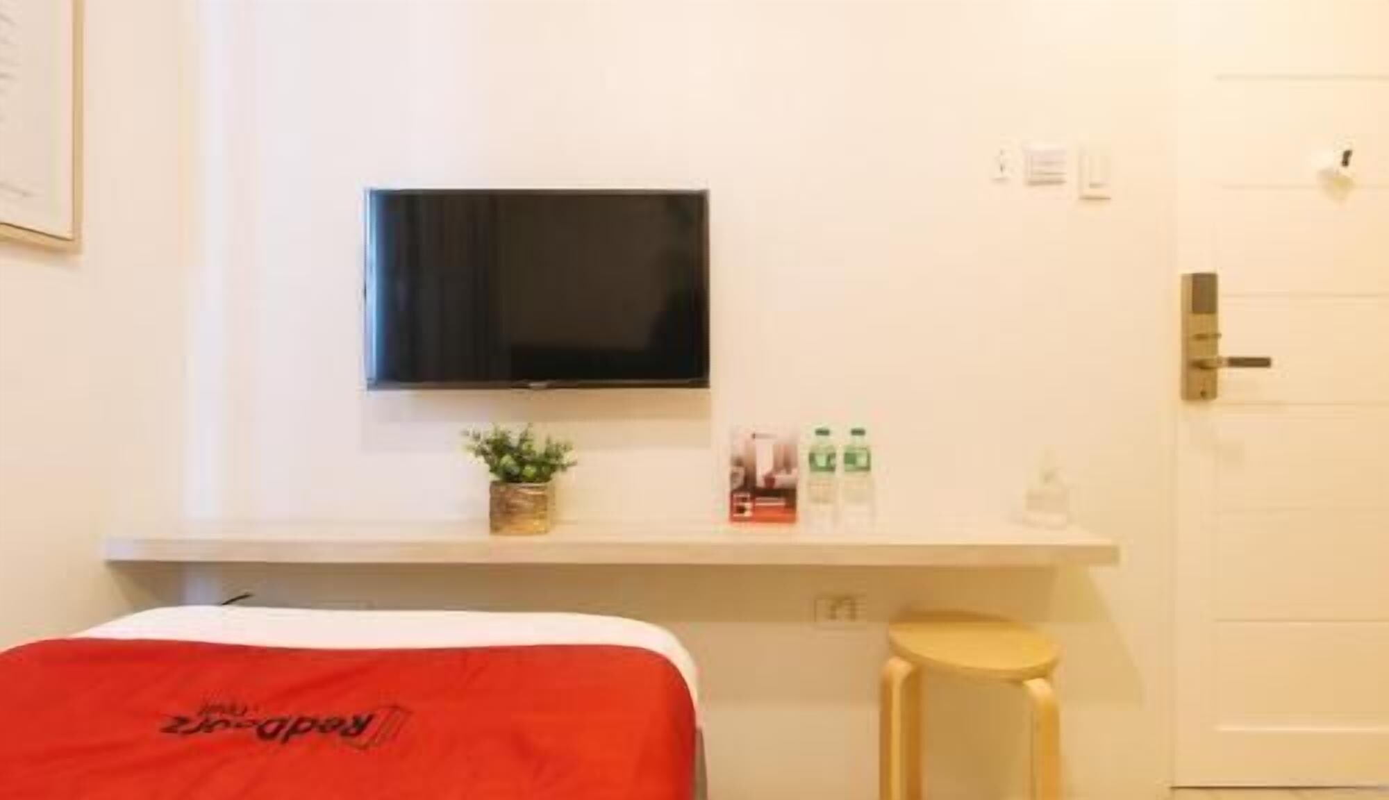 A hotel room with a bed, a small stool, a TV mounted on the wall, and a door.