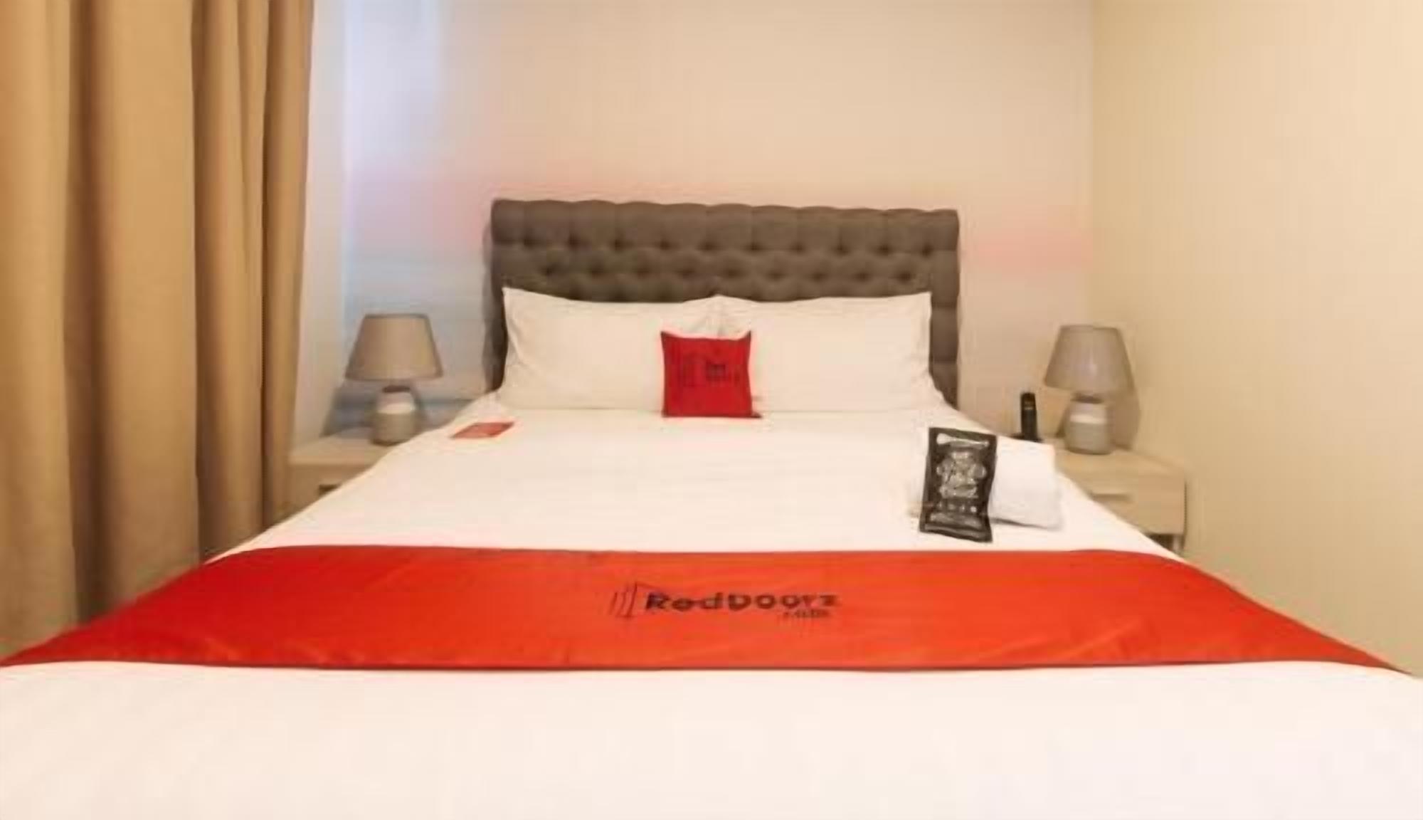 A hotel room with a bed, a red throw, a pillow, a nightstand, and a lamp.