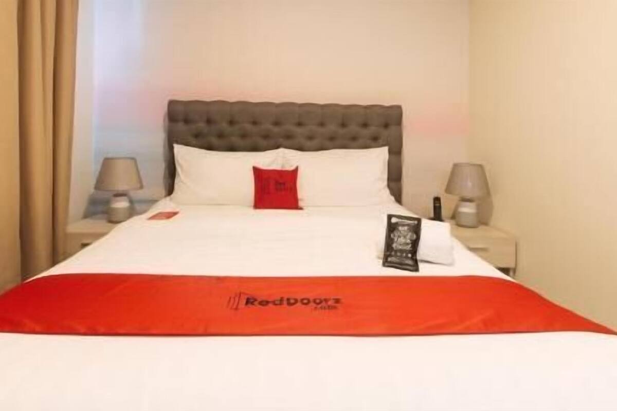 Une chambre d’hôtel avec un lit, un plaid rouge, un oreiller, une table de chevet et une lampe.