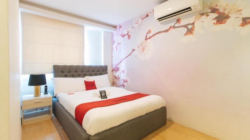A bedroom with a bed, a nightstand, a lamp, a wall-mounted air conditioner, and a window.