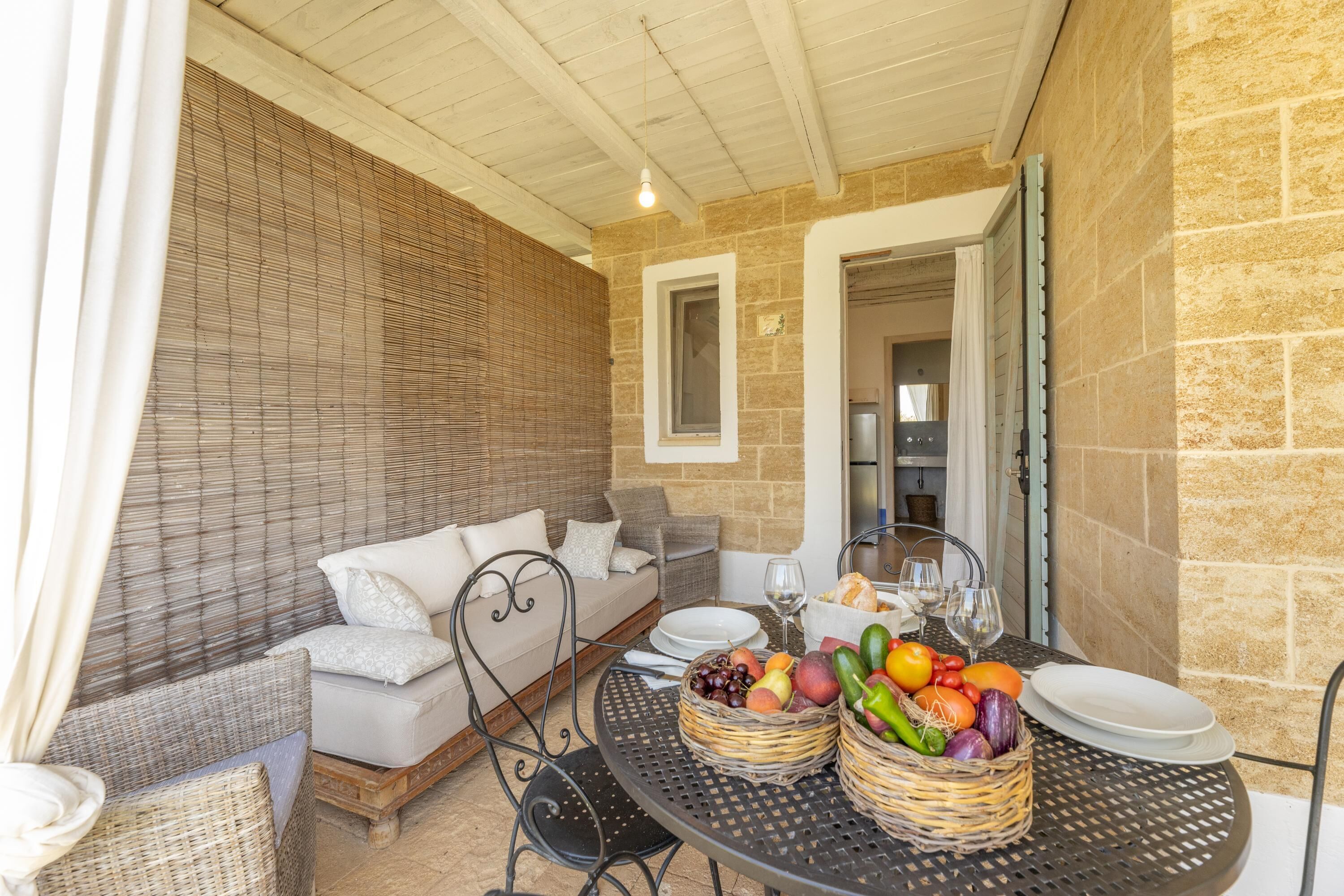 A cozy outdoor seating area with wicker furniture, a round table with fruit baskets, and a stone wall backdrop.