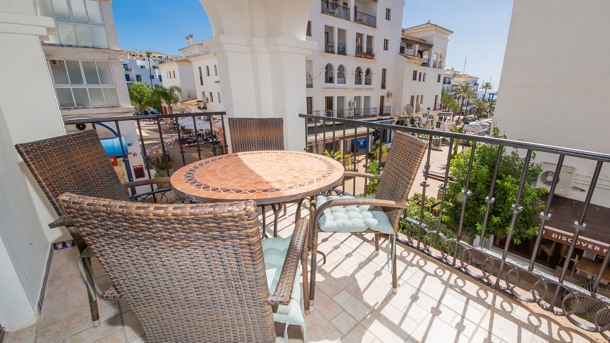 A balcony with wicker furniture, a tiled table, and a view of buildings and palm trees.