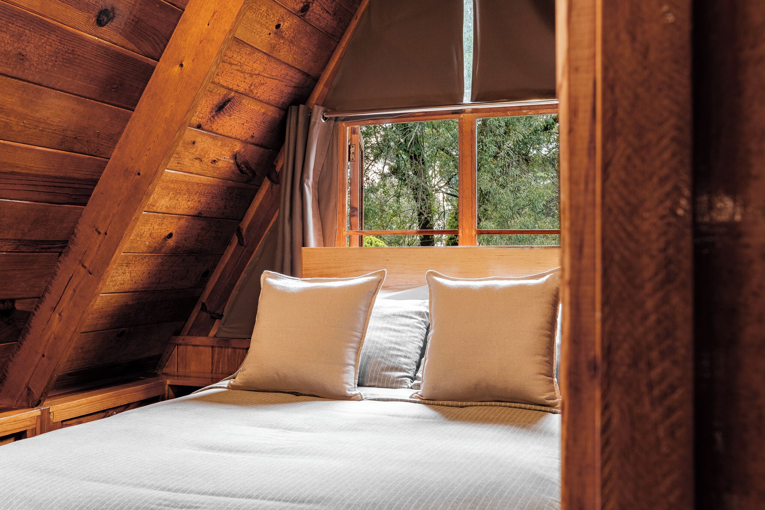 A bedroom with a sloped ceiling, a bed with two pillows, and a window with a view of trees.