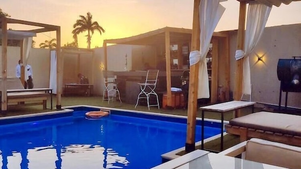 A swimming pool with lounge chairs and a covered seating area, surrounded by simple structures and palm trees.