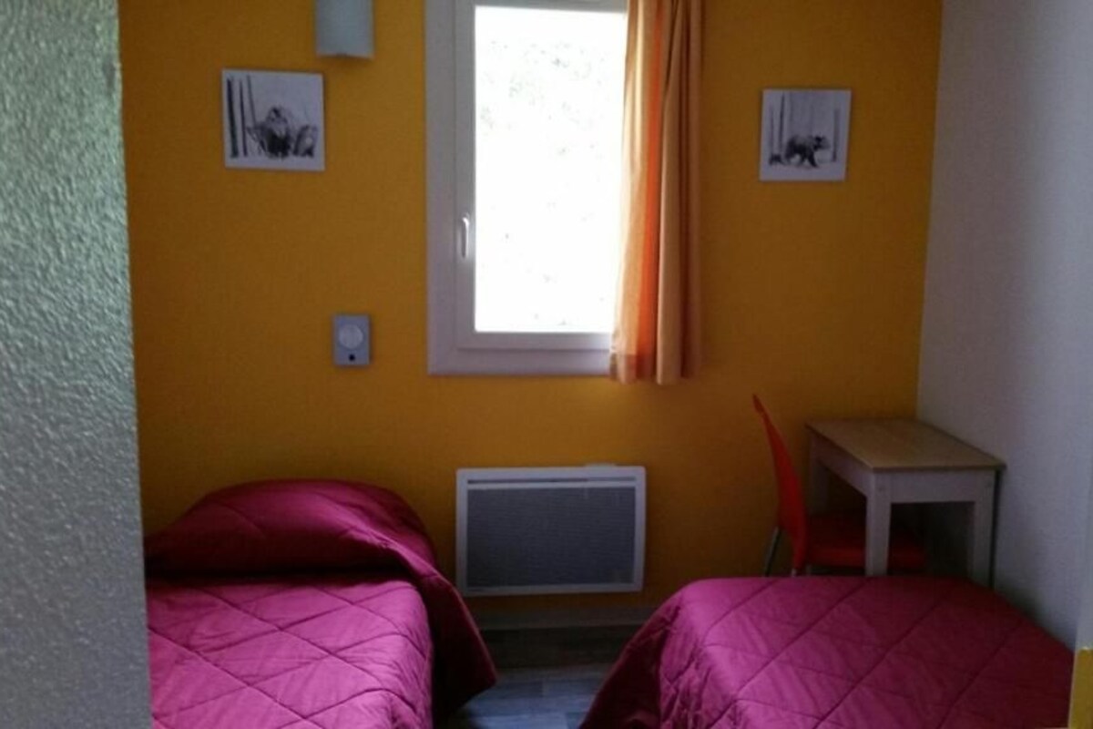 A small room with two beds, a window with curtains, a radiator, and a small table.