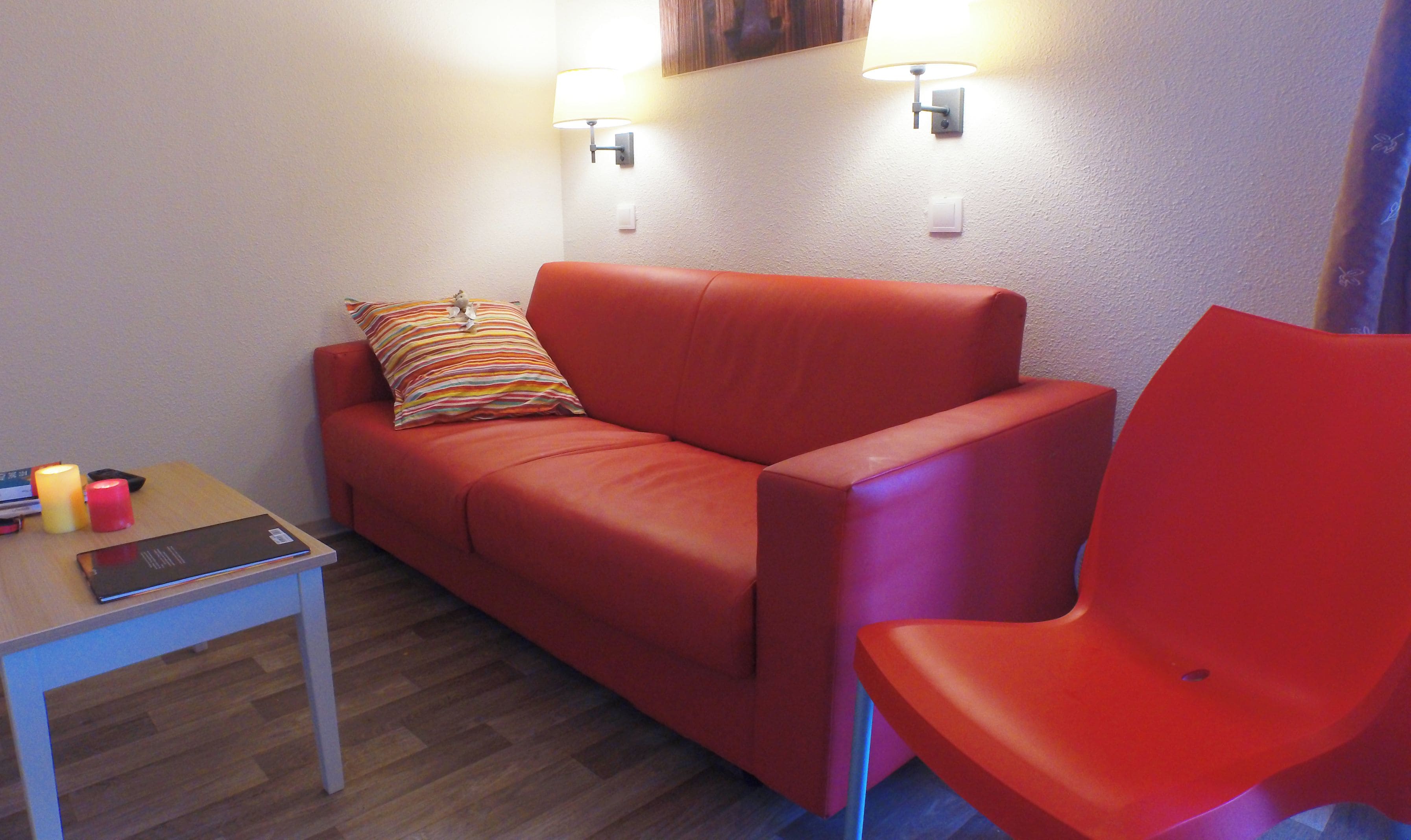 Un canapé rouge avec un coussin à rayures, un fauteuil rouge, une petite table avec un livre et des bougies, et deux lampes fixées au mur.