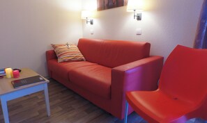 En rød sofa med en stribet pude, en rød lænestol, et lille bord med en bog og stearinlys, samt to vægmonterede lamper.