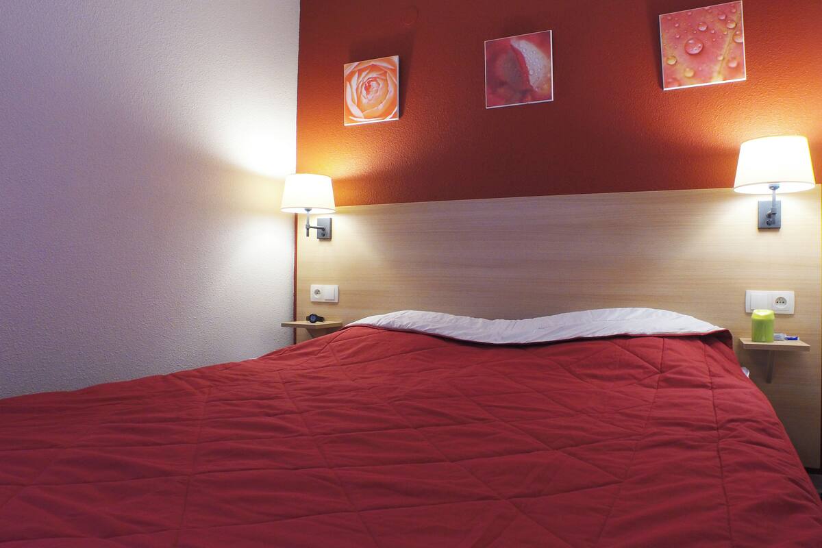 Een hotelkamer met een rode sprei, twee bedlampen en drie ingelijste foto’s aan de muur.