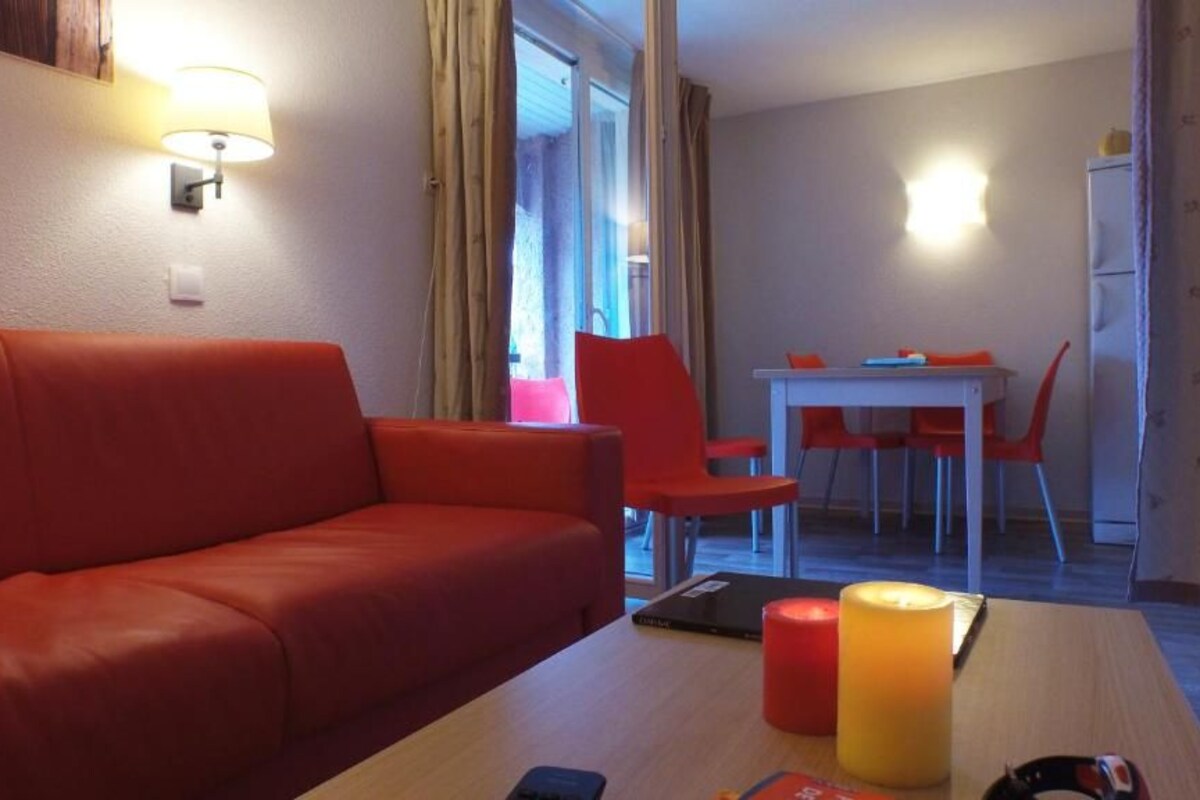 Um quarto com um sofá vermelho, uma mesa de madeira com uma vela e uma área de jantar com cadeiras vermelhas.