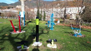 Een buitenoefenzone met diverse fitnessapparaten, waaronder een rode en groene paal, een blauwe machine en een gele constructie.