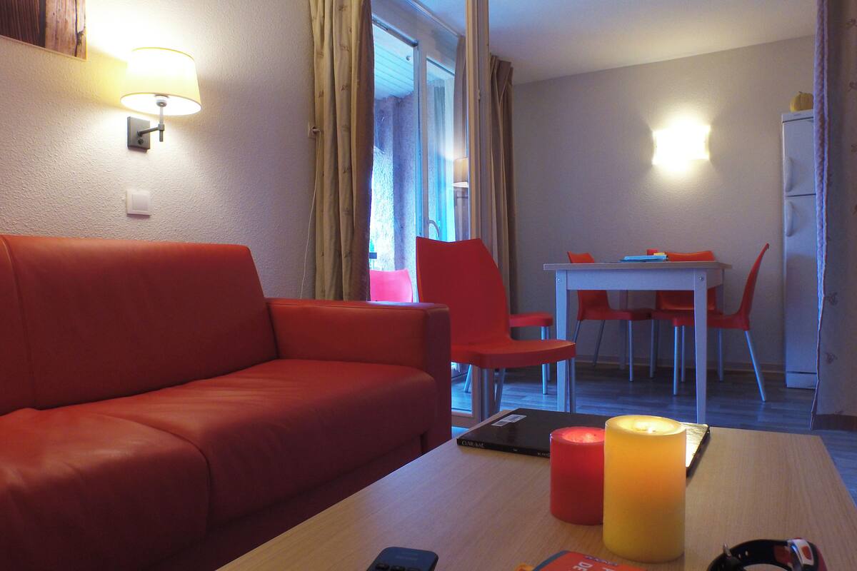 Een hotelkamer met een rode bank, een houten tafel met kaarsen, een afstandsbediening en een eethoek met een witte tafel en rode stoelen.