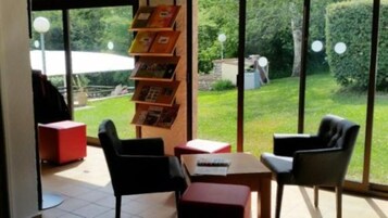 Quarto com uma janela grande, estante de livros e área de estar com cadeiras e mesa.