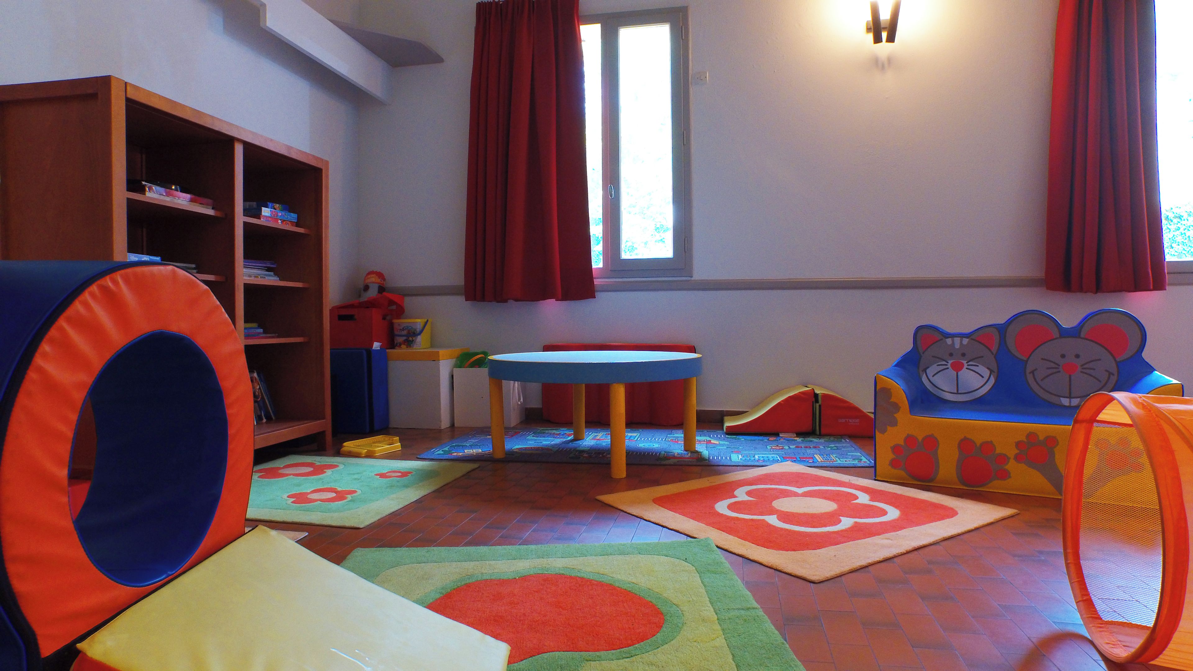 A children's playroom with a bookshelf, a table, and various play mats.