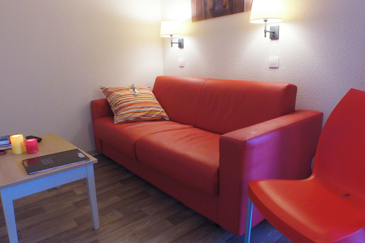 A red sofa with a striped pillow, a red chair, a small table with a book and candles, and two wall-mounted lamps.