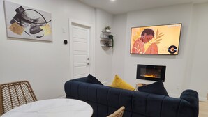 A living room with a dark blue sofa, a round white table, wicker chairs, a white door, a wall-mounted TV, and a fireplace. - Deluxe 2-BR Basement Suite in South East Calgary (Calgary)