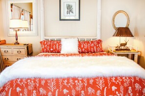 A bed with a white and red floral bedspread, a white fluffy pillow, and two red pillows with a white floral pattern. There is a wooden nightstand with a lamp, a framed picture on the wall, and a mirror above the nightstand. - EML 9 Juniper (Paonia)