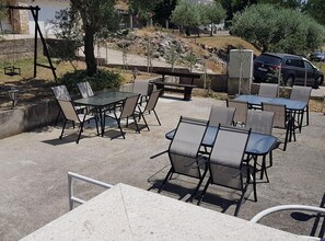 An outdoor patio area with multiple tables and chairs, a stone wall, and a parked car in the background. - Apartments Danijela & Vlado (Lopar)