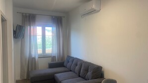 Una sala pequeña con un sofá seccional gris, un televisor de pantalla plana en la pared y una ventana con cortinas.
