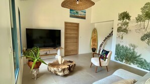 Un salon moderne doté d’un grand téléviseur à écran plat, d’un plafond en bois, d’une fresque murale sur le thème de la plage, d’une planche de surf et d’un canapé confortable.