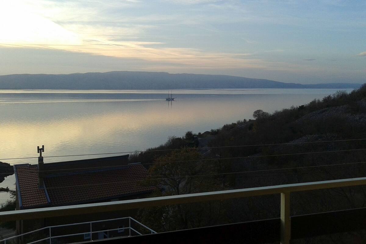 A view of a calm lake with a distant boat, surrounded by hills and a residential area with tiled roofs.