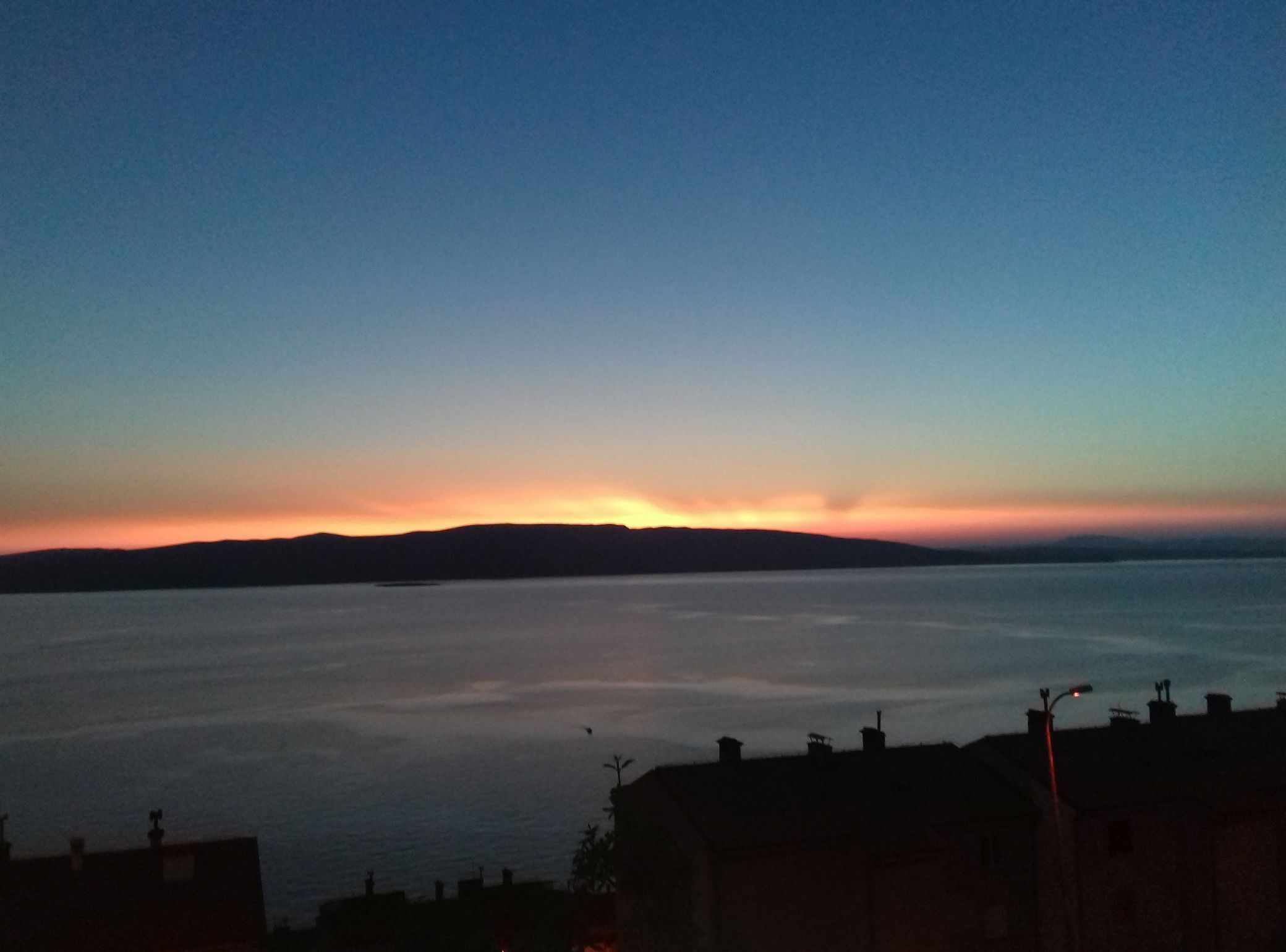 Paisagem costeira com um pôr do sol sobre a água, formas de relevo em silhueta e edifícios residenciais ao primeiro plano.
