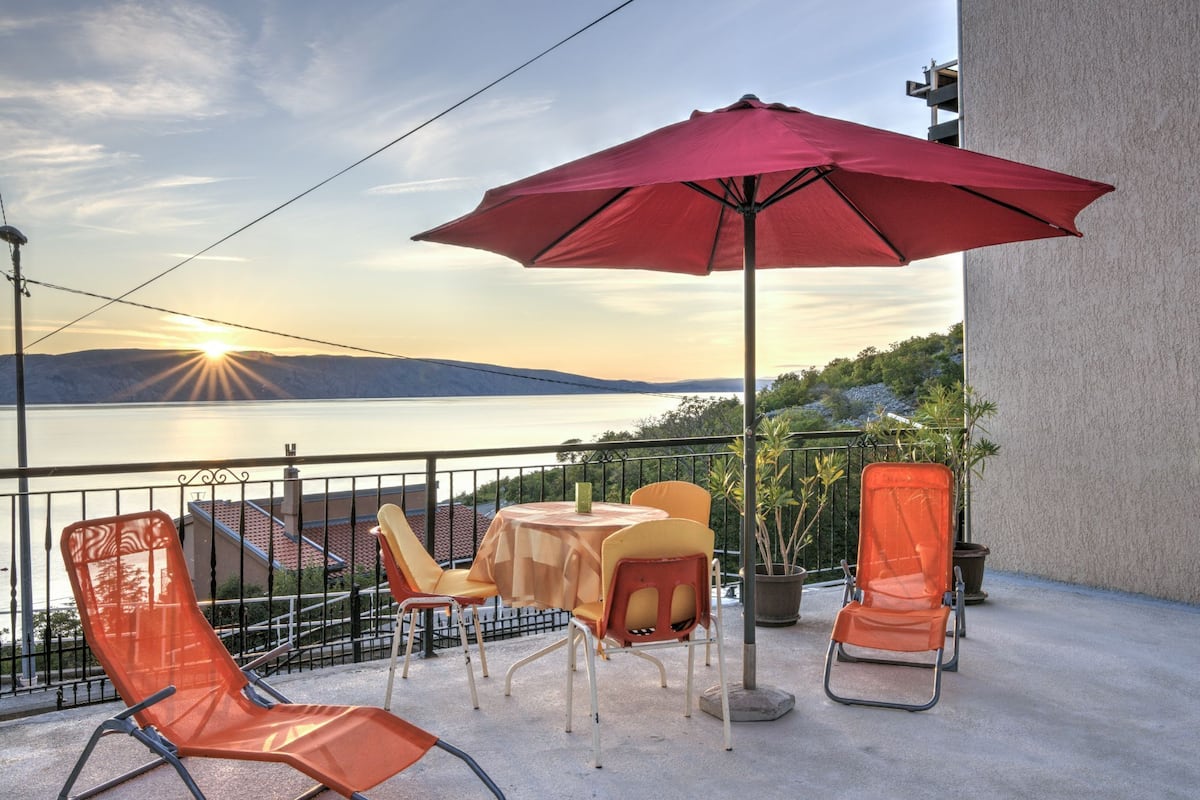 A balcony with a table, chairs, and a red umbrella overlooking a body of water and hills.