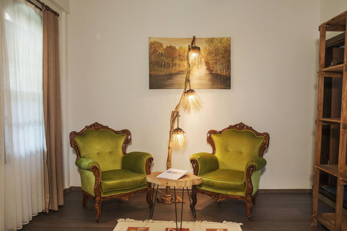 A room with two green upholstered chairs, a wooden floor, a painting on the wall, and a wooden bookshelf.