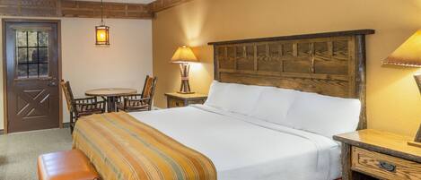 Una habitación de hotel con cabecera de madera, cama con ropa blanca, una banca al pie de la cama, mesita de noche de madera, una mesa con sillas y una lámpara.