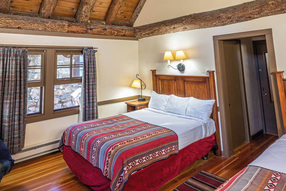 A bedroom with a wooden ceiling, a bed with a patterned bedspread, a nightstand with a lamp, and a window with curtains.