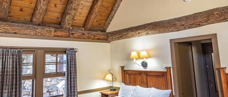 A bedroom with a wooden ceiling, a bed with a patterned bedspread, a nightstand with a lamp, and a window with curtains.