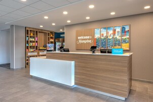 Zona de recepción de hotel con diseño moderno, que incluye un mostrador de recepción, un cartel de 'Welcome to Hampton', una obra de arte con un paisaje urbano y un cartel de 'Hilton Honors'.
