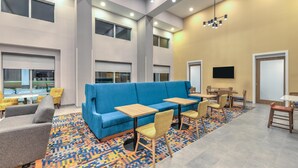 A modern lounge area with a blue sofa, wooden tables, and chairs, a patterned rug, and large windows. - Hampton Inn & Suites Little Rock Airport (Little Rock)