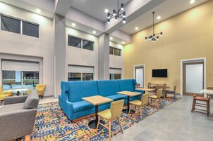 Un espacio lounge moderno con un sofá azul, mesas y sillas de madera, un tapete estampado y ventanas amplias.