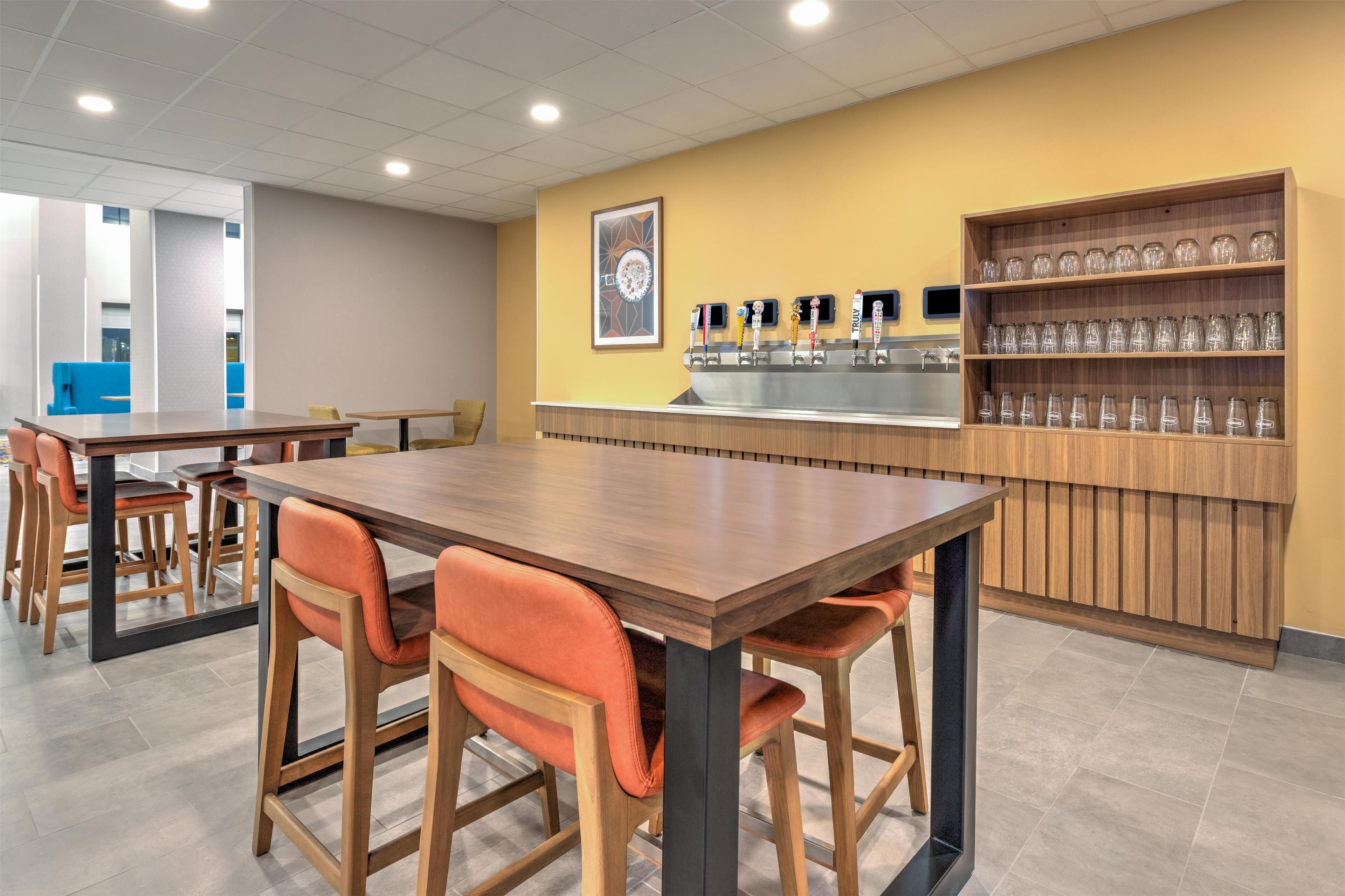Un café moderno con mesas y sillas de madera, una barra con máquina de café y un estante con vasos.