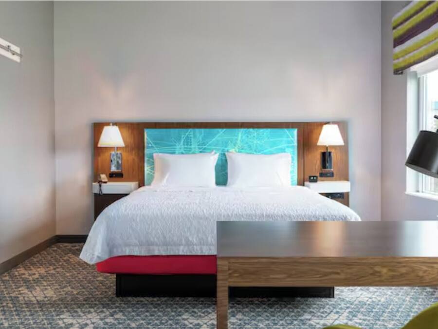 A hotel room with a large bed, two bedside lamps, a wooden headboard, a window with a view, and a small table.