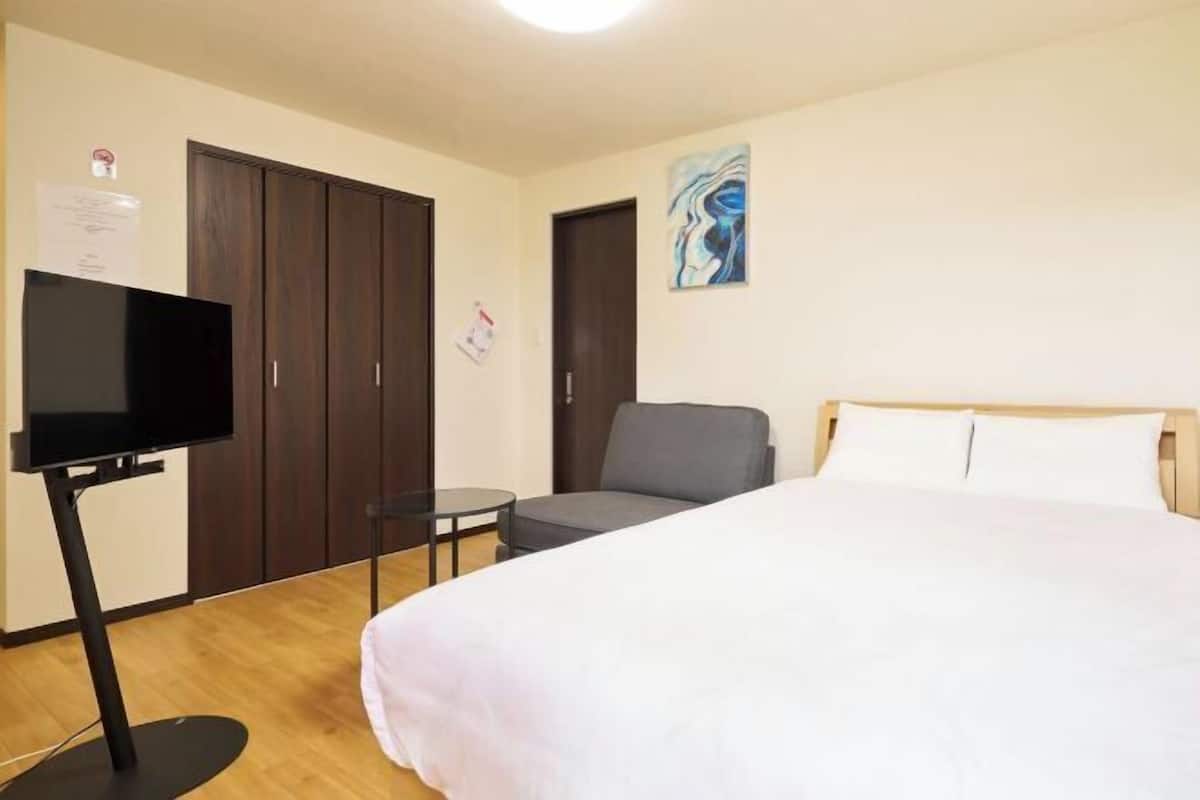 A hotel room with a bed, a TV, a chair, a small table, and a wardrobe.