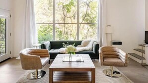 A living room with a sofa, two armchairs, a coffee table, and a large window with curtains. - Treehouse studio, secluded & chic hiker’s paradise

 (San Anselmo)