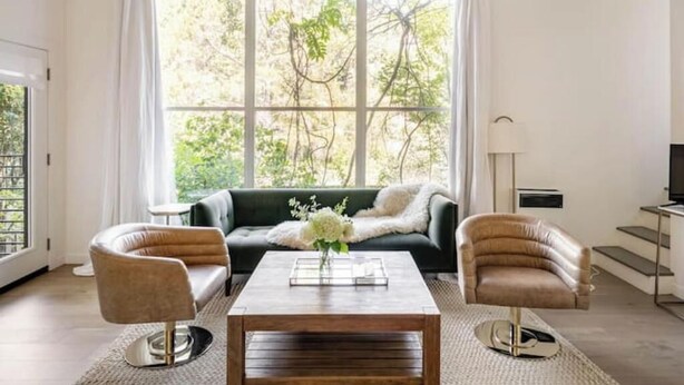 A living room with a sofa, two armchairs, a coffee table, and a large window with curtains. - Treehouse studio, secluded & chic hiker’s paradise

 (San Anselmo)