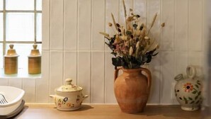 A kitchen with a wooden countertop, a white kettle, a vase with dried flowers, and a framed painting above the counter.