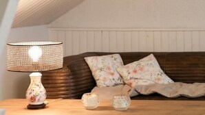 A cozy room with a wooden table, a lamp, a sofa with floral cushions, and a framed picture on the wall.