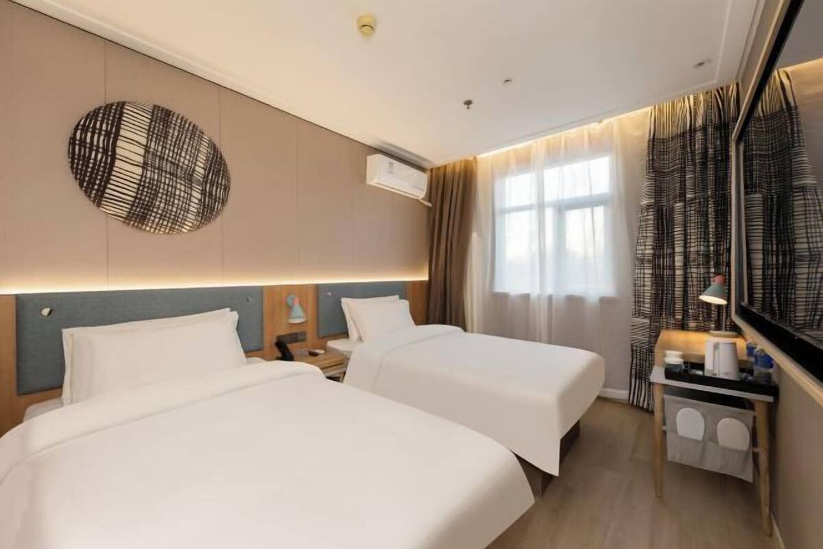 A hotel room with two beds, a bedside table with a lamp, an air conditioning unit, and a large mirror with curtains.