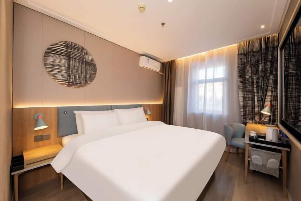 A modern hotel room with a large bed, a desk, a chair, and a wall-mounted air conditioner.