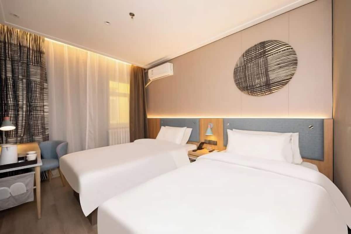 A hotel room with two beds, a desk, a chair, a lamp, and a wall art piece.