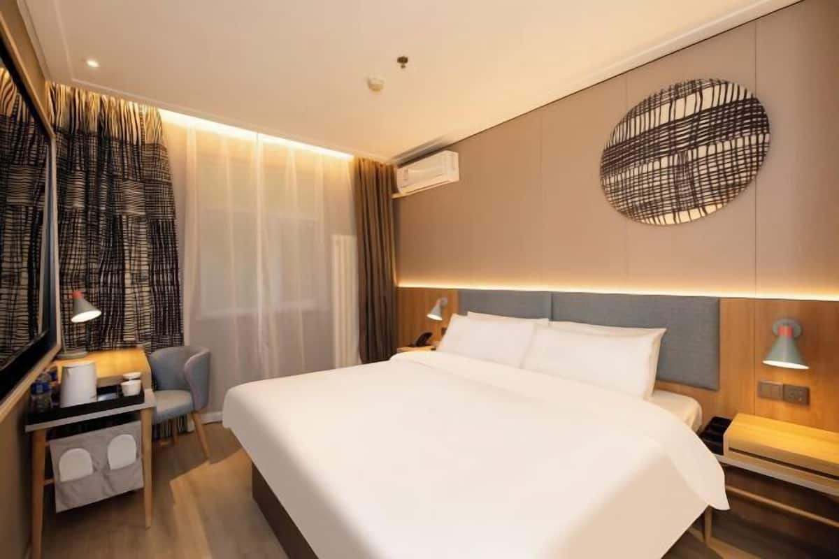 A modern hotel room with a large bed, a desk with a chair, a wall-mounted air conditioner, and a decorative circular wall piece.